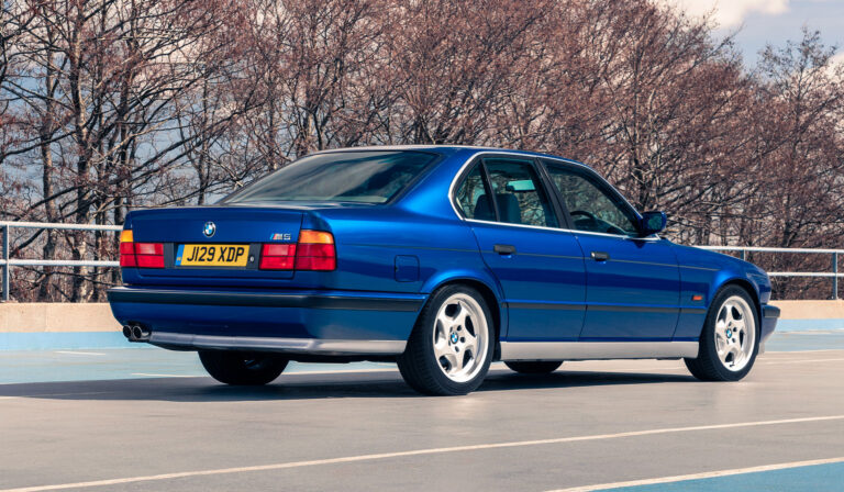 Tumesinine BMW E34 M5 tagant külgvaates, pargitud avarale asfalteeritud platsile, taustal raagus puud.