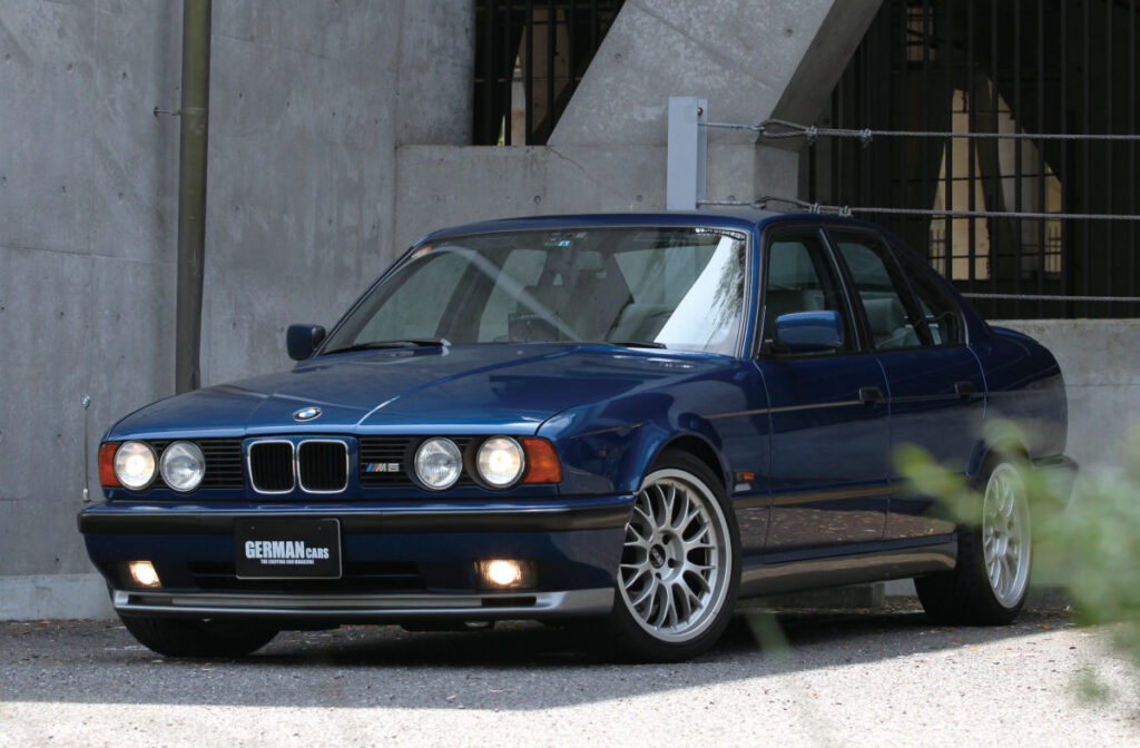 BMW M5 (E34) Tumesinine BMW E34 M5 esiosas ja külgvaates, pargitud betoonseina taustal, sisselülitatud tuledega.