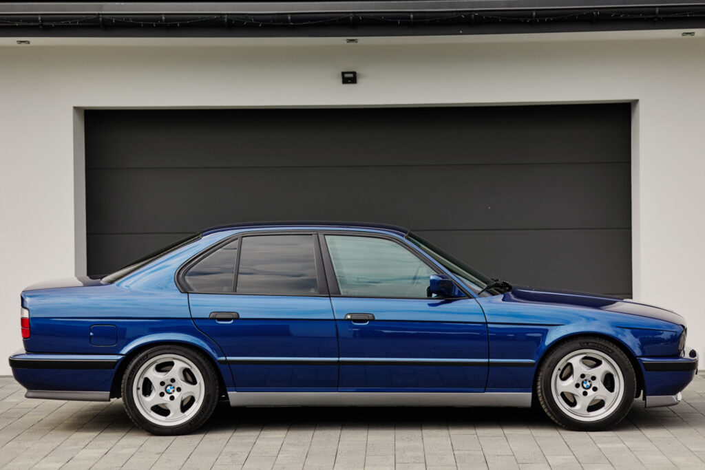Tumesinine BMW M5 (E34) külgvaade Tumesinine BMW M5 (E34) Nürgburg Pack külgvaates, pargitud kaasaegse maja tumeda garaažiukse ette.