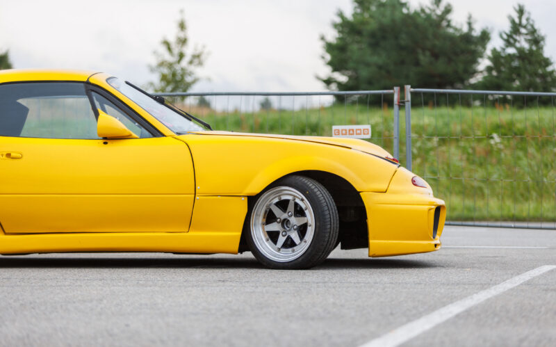 Kollane madaldatud Mazda MX-5 NA sportauto laiendatud poritiibade ja poleeritud randiga retrovelgedega Porsche Ringi asfaltplatsil Augustfest 2026 festivalil