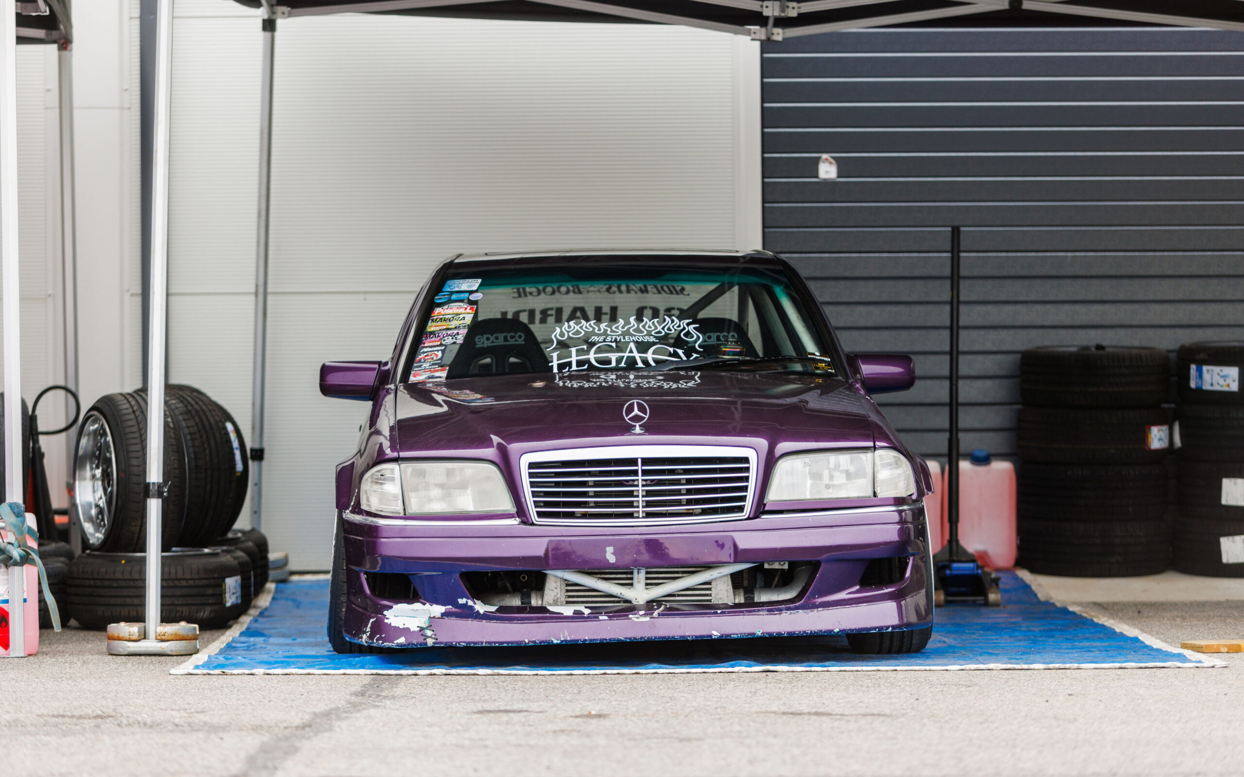 Lilla Mercedes-Benz C-klassi W202 driftiauto suure vahejahuti ja Sparco istmetega hooldustelgi all Porsche Ringi boksiteel Augustfest 2026 festivalil