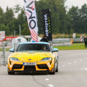 Eestvaates kollase-sinise disainkilega Toyota GR Supra sportauto sõitmas Porsche Ringi boksiteel Monster Energy lipu taustal Augustfest 2026 festivalil