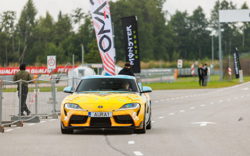 Eestvaates kollase-sinise disainkilega Toyota GR Supra sportauto sõitmas Porsche Ringi boksiteel Monster Energy lipu taustal Augustfest 2026 festivalil