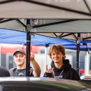 Naeratavad noored festivalikülastajad ja tiimiliikmed telgi all käemärkidega tervitamas Porsche Ringi boksiteel Augustfest 2026 festivalil