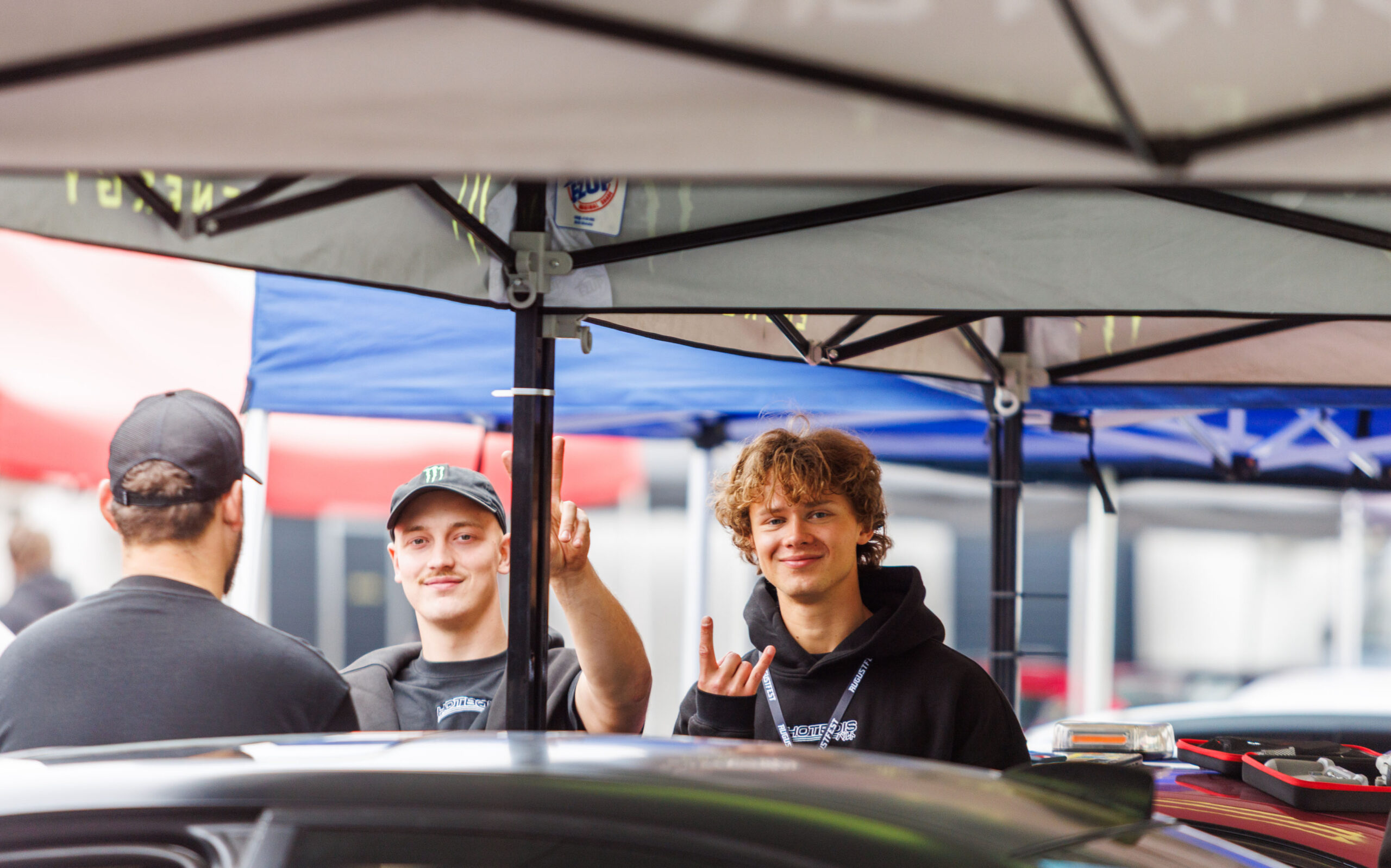Naeratavad noored festivalikülastajad ja tiimiliikmed telgi all käemärkidega tervitamas Porsche Ringi boksiteel Augustfest 2026 festivalil