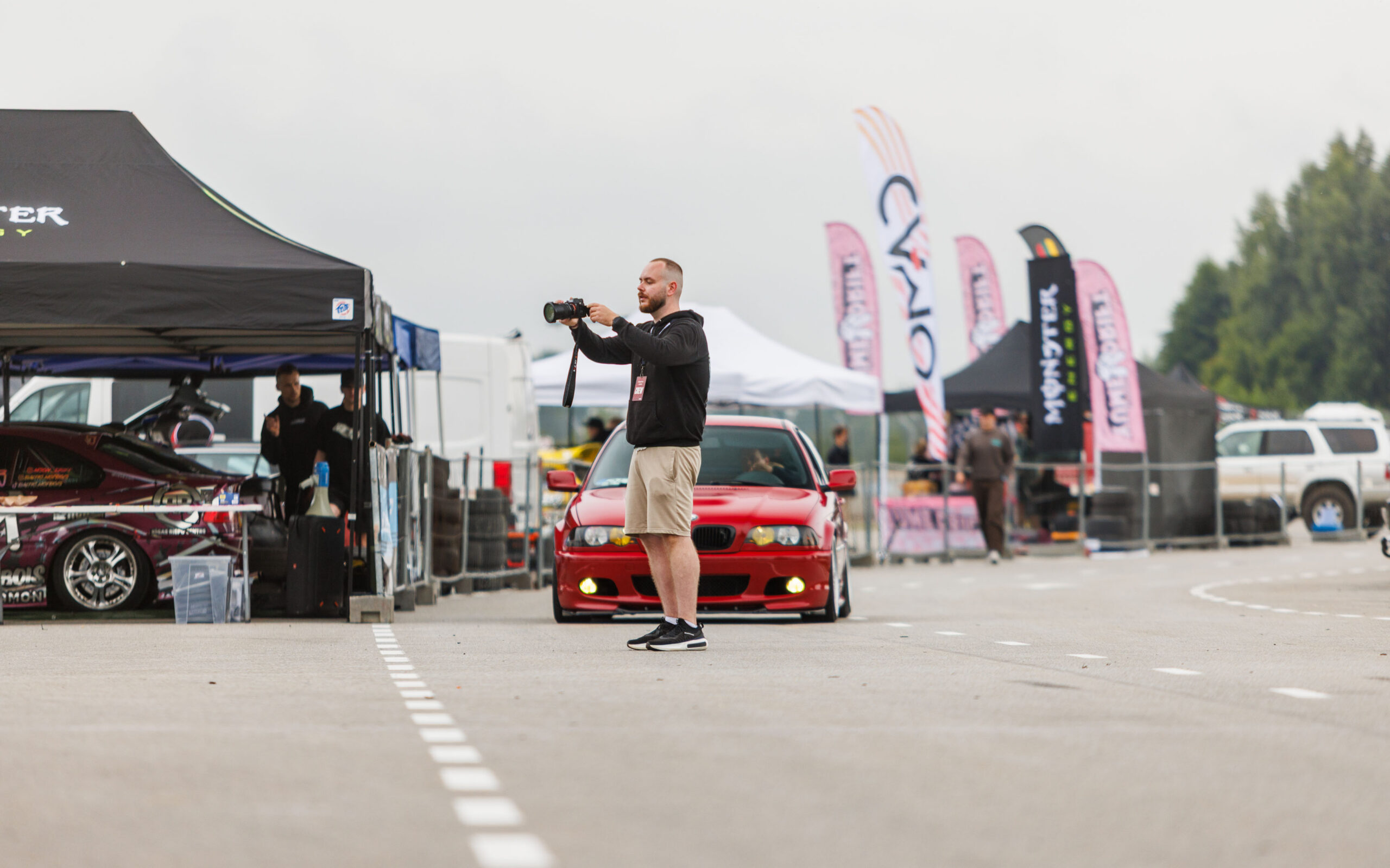 Fotograaf kaameraga pildistamas punast madaldatud BMW E46 sedaani Porsche Ringi boksiteel Monster Energy telkide ees Augustfest 2026 festivalil