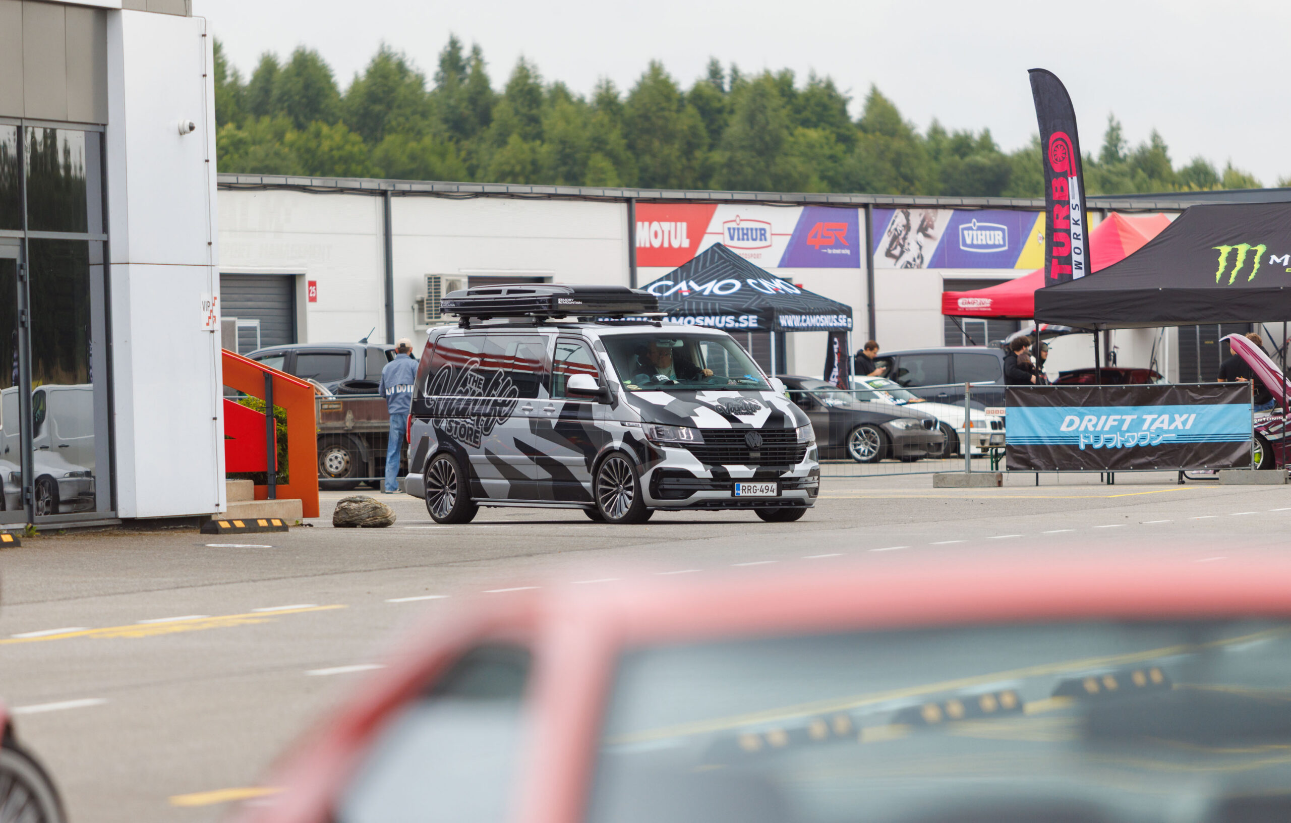 Musta-halli kamoflaažkilega kaetud ja katuseboksiga Volkswagen Transporter T6 buss liikumas Porsche Ringi Drift Taxi ala ees Augustfest 2026 festivalil