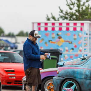 Mees nokamütsis puhastamas lapiga kirjut värvi Mazda MX-3 tuuningautot punase klapptuledega sportauto ees Pärnu Porsche Ringi alal Augustfest 2026 festivalil