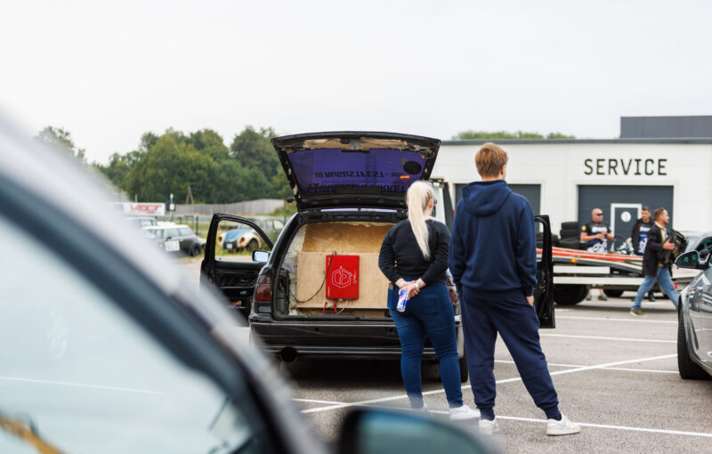 Musta universaalkerega auto avatud tagaluuk kohandatud helisüsteemi vineerkasti ja punase võimendiga, ees seismas kaks pealtvaatajat Porsche Ringi teenindushoone ees Augustfest 2026 festivalil