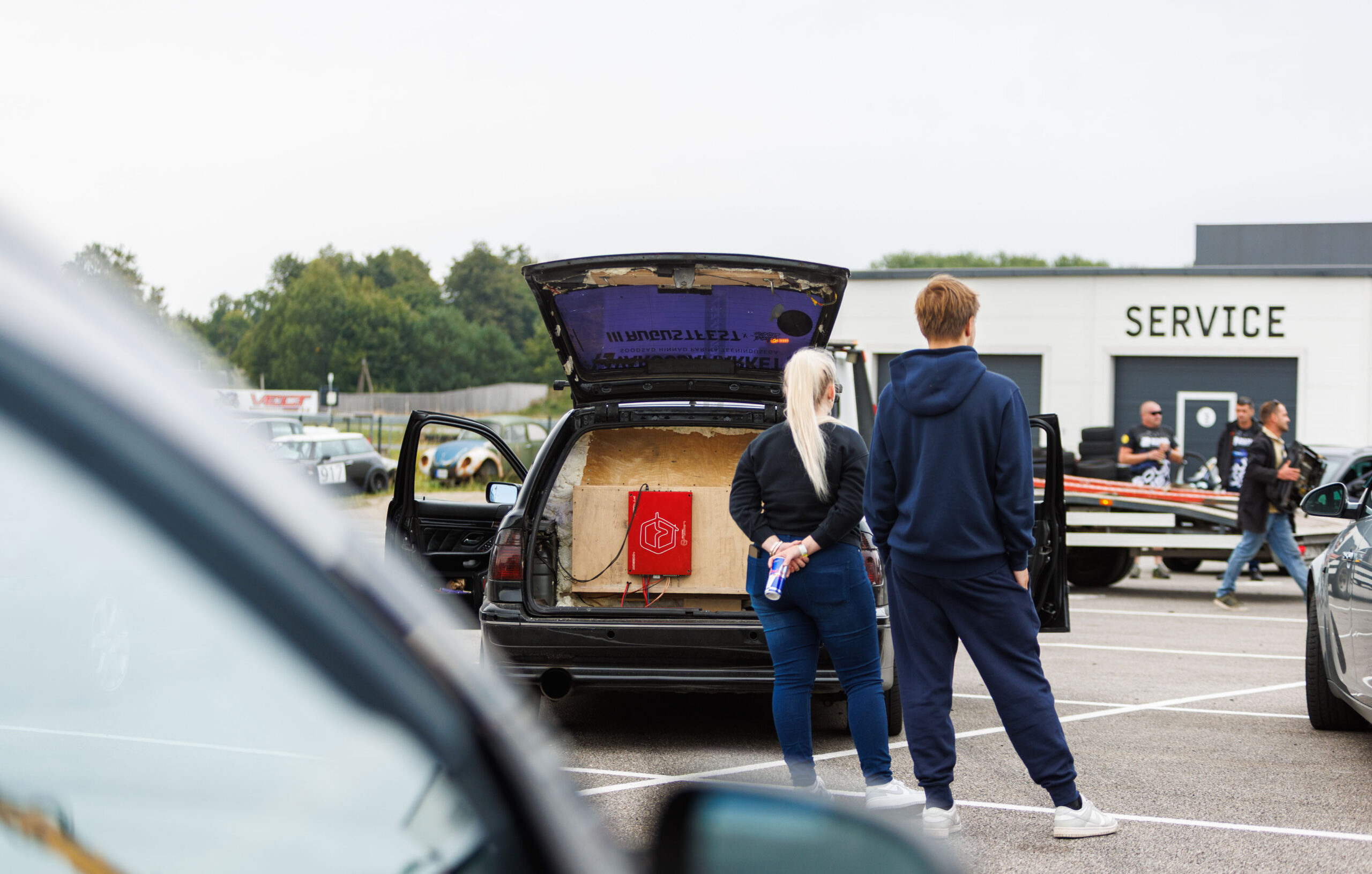 Musta universaalkerega auto avatud tagaluuk kohandatud helisüsteemi vineerkasti ja punase võimendiga, ees seismas kaks pealtvaatajat Porsche Ringi teenindushoone ees Augustfest 2026 festivalil