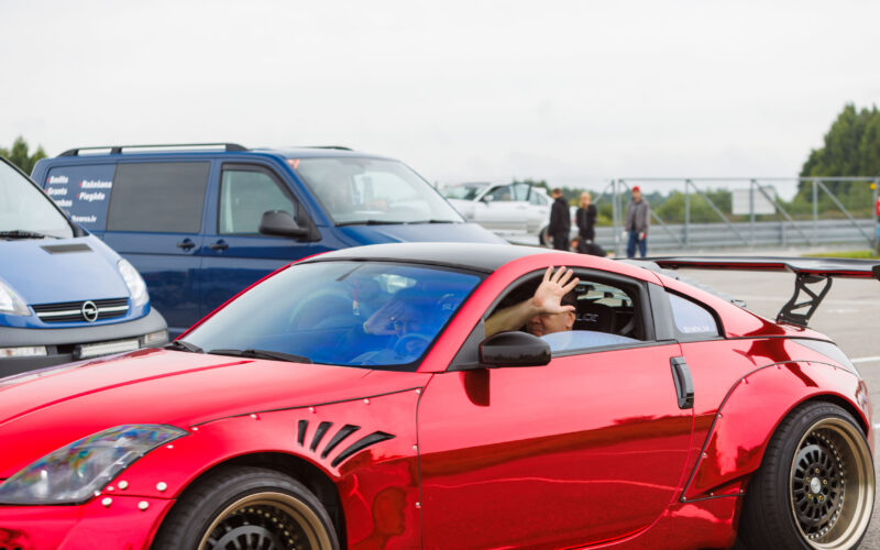 Punane kroomitud widebody Nissan 350Z driftiauto suure tagatiiva ja laiade velgedega, mille juht lehvitab aknast Porsche Ringil Augustfest 2026 festivalil