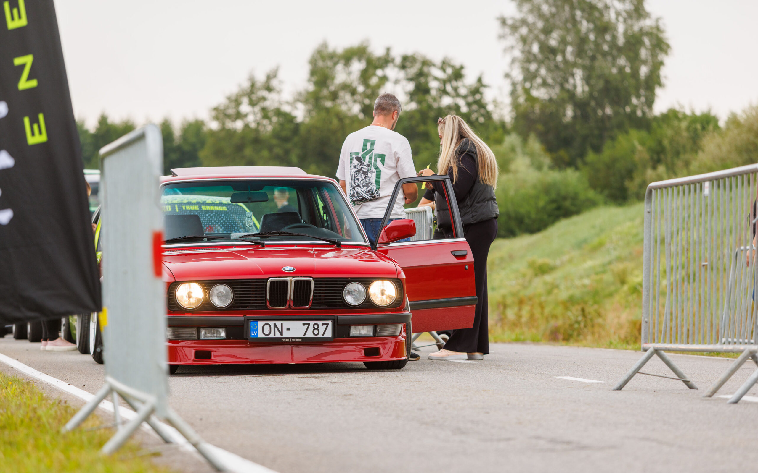 Punane madaldatud BMW E30 sedaan avatud ukse ja põlevate tuledega sissesõiduväravas Pärnu Porsche Ringil Augustfest 2026 festivalil