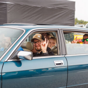 Rõõmsad inimesed sinakasrohelises klassikalises Jaguar XJ40 sedaanis käemärke näitamas Pärnu Porsche Ringil Augustfest 2026 festivalil