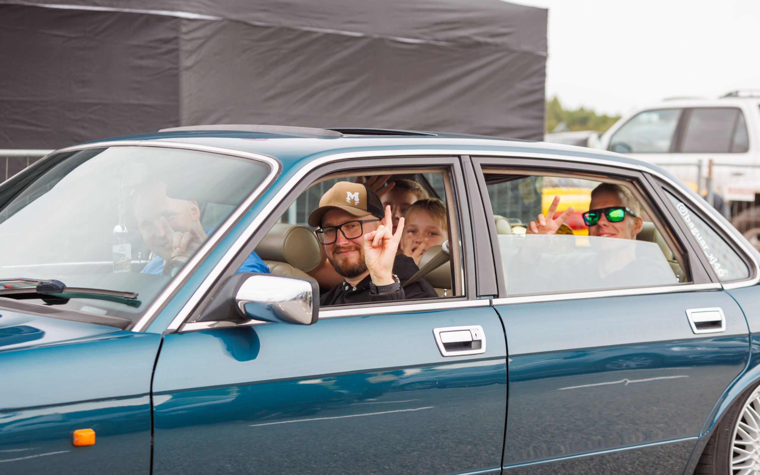 Rõõmsad inimesed sinakasrohelises klassikalises Jaguar XJ40 sedaanis käemärke näitamas Pärnu Porsche Ringil Augustfest 2026 festivalil