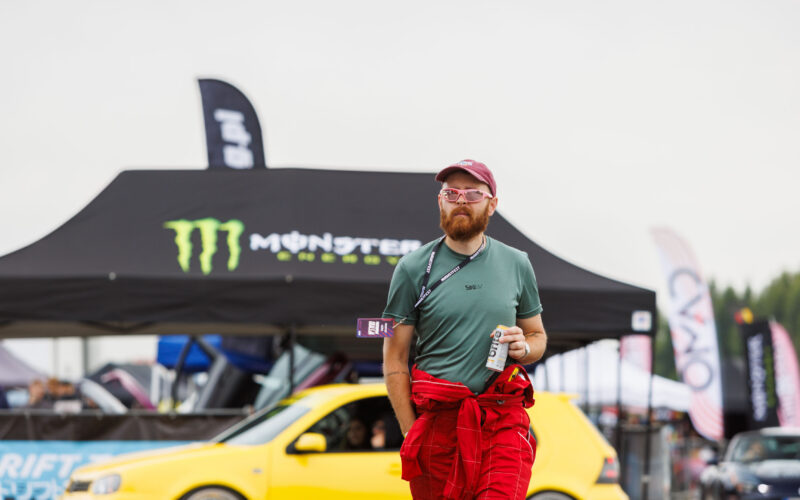 Punases võidusõidukombinesoonis ja päikeseprillidega mees kõndimas Monster Energy telgi ja kollase auto ees Pärnu Porsche Ringil Augustfest 2026 festivalil