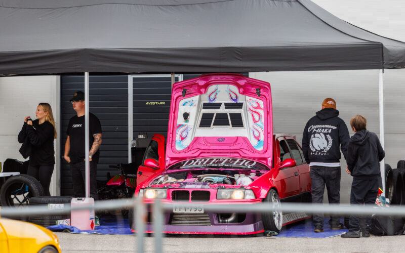 Erkroosa BMW E36 driftiauto avatud leegimustriga kapotiga ja Drift HQ kirjaga boksi telgi all Pärnu Porsche Ringil Augustfest 2026 festivalil